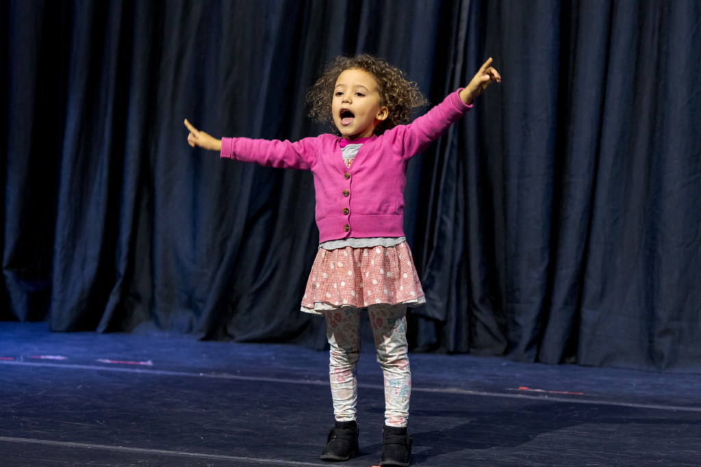A child actress on stage in front of a curtain.